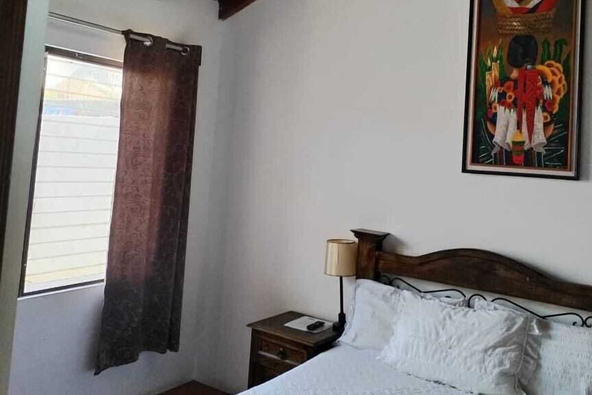 Un dormitorio con techo de madera, una cama con ropa blanca, una mesita de noche, una lámpara, una ventana con cortinas y un cuadro en la pared.
