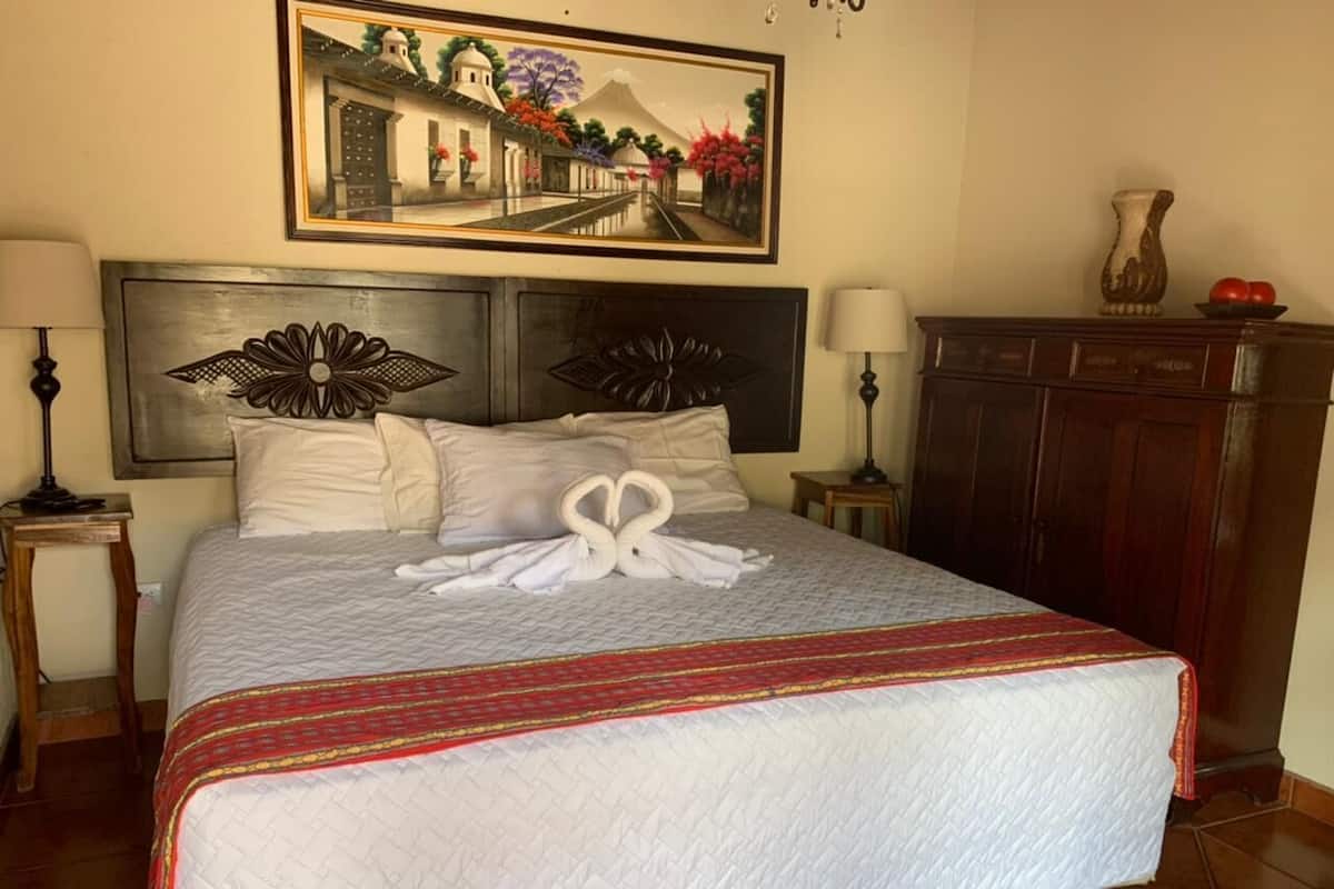 Una cama bien tendida con una toalla dispuesta en forma de corazón, un cuadro encima y un aparador de madera a la derecha.