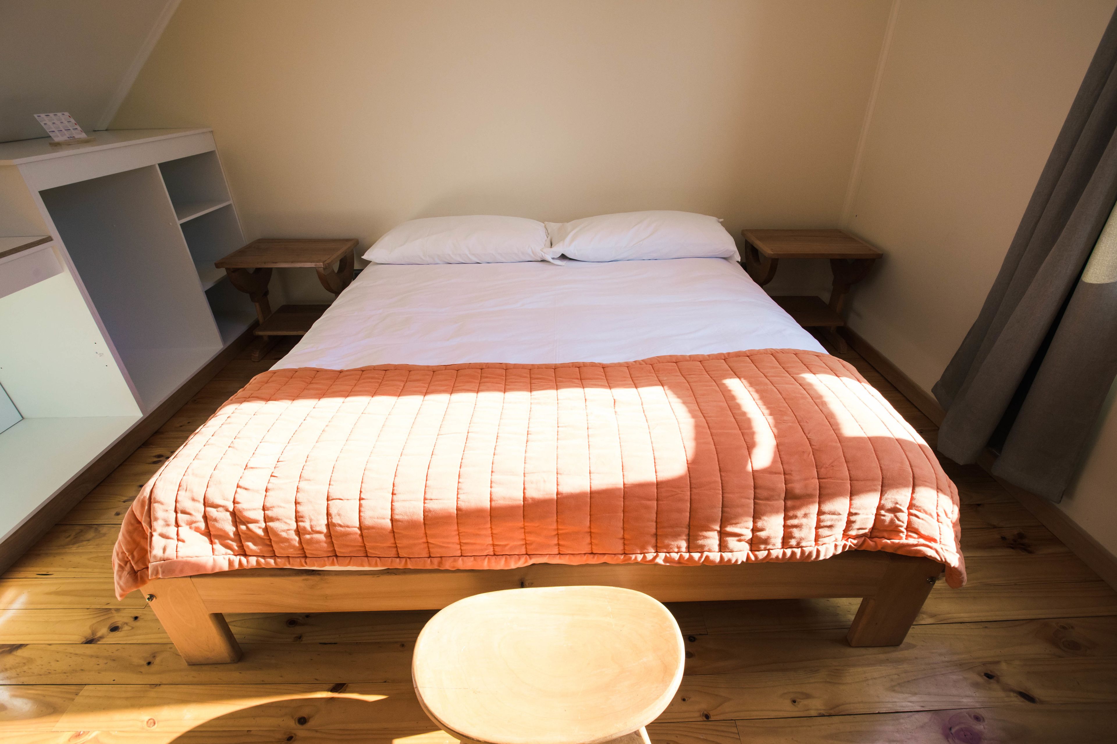 A neatly made bed with a wooden frame and a pink quilt, two bedside tables, and a small wooden stool.