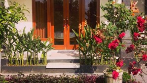 A two-story house with a tiled roof, a porch with a hanging light fixture, and a variety of plants and flowers in the front yard. - Pondok Ayu Homestay (Ubud)