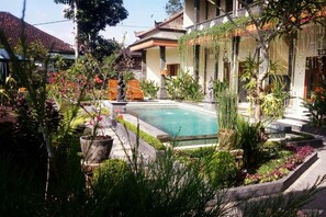A pool surrounded by lush greenery and a traditional building with a tiled roof. - Pondok Ayu Homestay (Ubud)