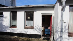 Un pequeño edificio de una sola planta con fachada blanca, una puerta que da a una habitación con cortina roja y una ventana con marco marrón.
