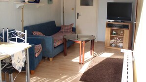 Een woonkamer met een blauwe bank, een glazen salontafel, een televisie op een houten dressoir en een klein kastje met decoratieve objecten.