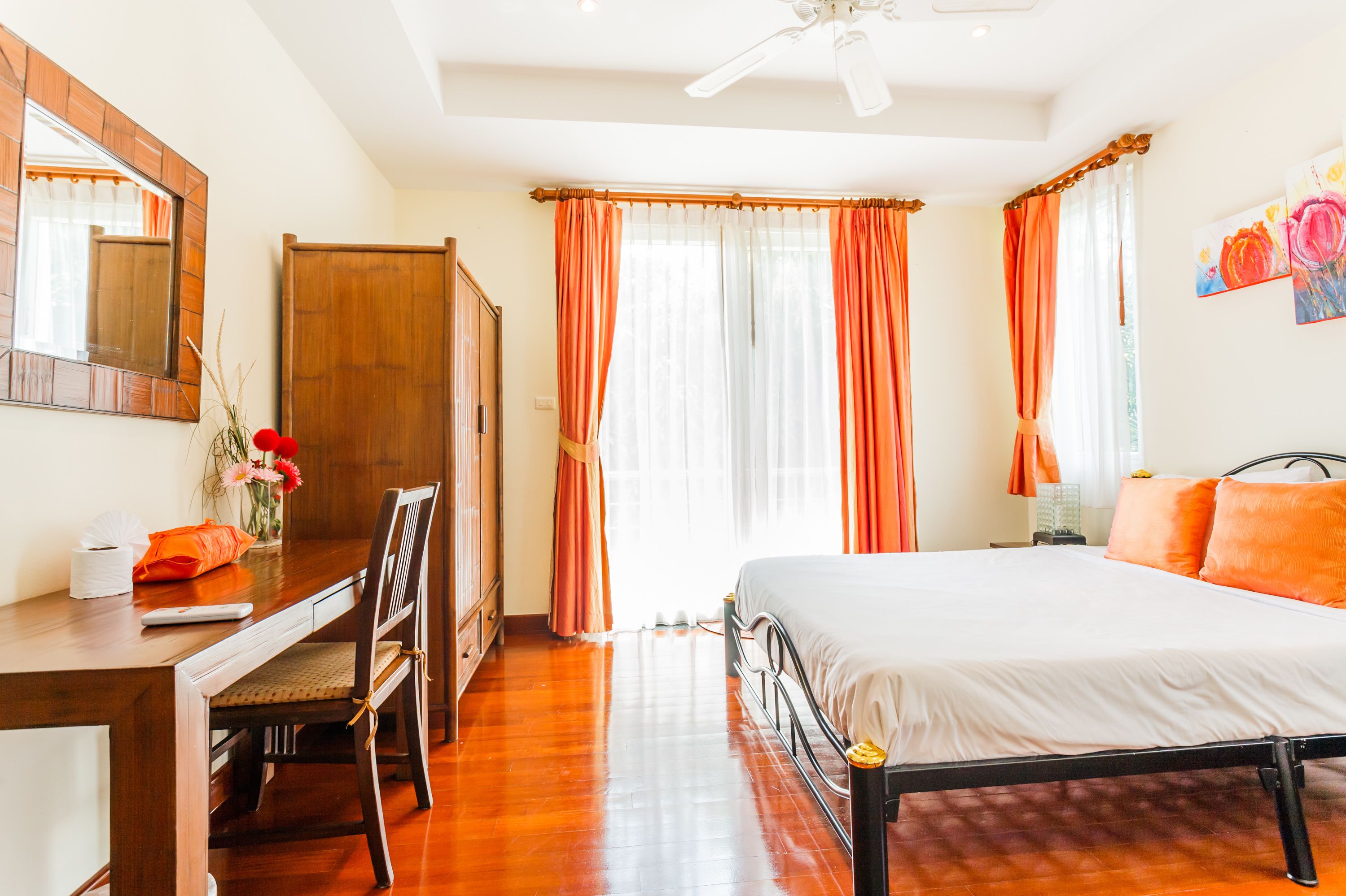 A bedroom with a wooden desk, a bed with orange bedding, a mirror, and a ceiling fan.