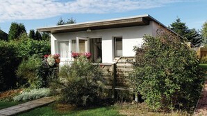 Une maison de plain-pied avec un toit plat, des murs extérieurs blancs et un petit porche avec des marches. Des plantes et des fleurs en pot sont disposées sur le porche, et la maison est entourée d’un jardin agrémenté de divers arbustes et arbres.