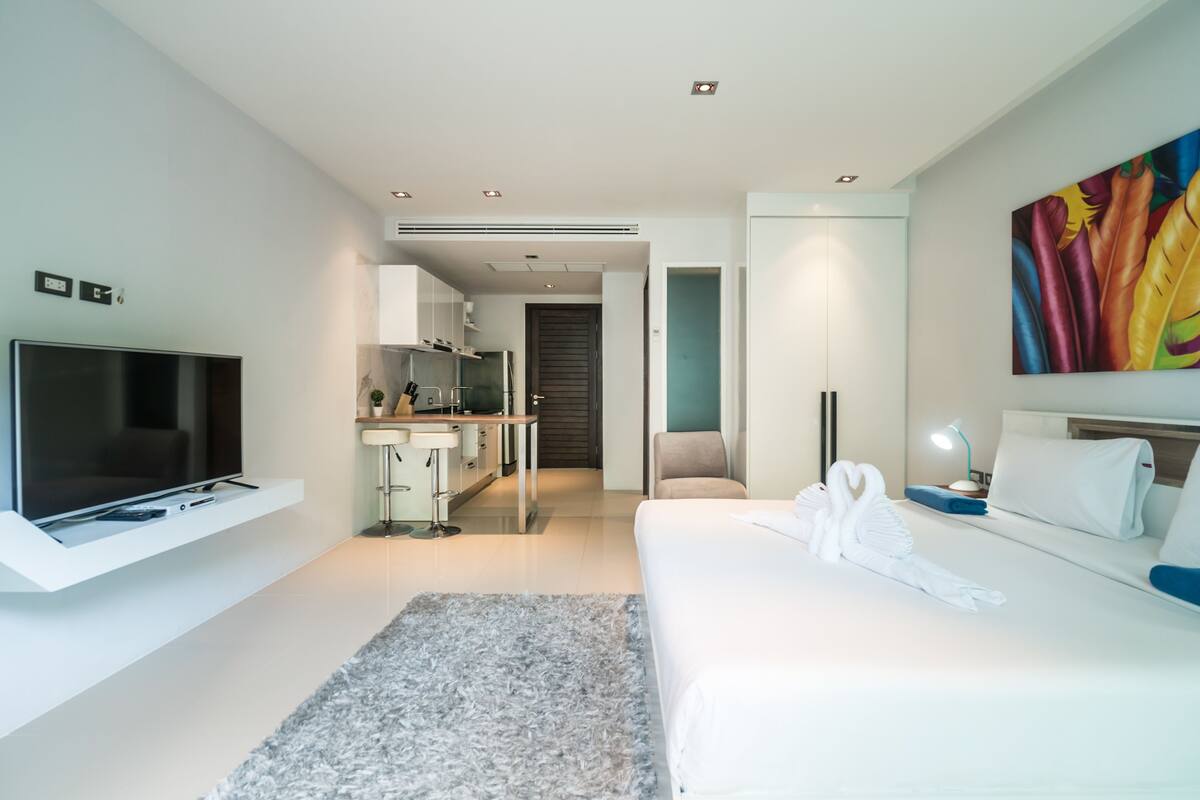 Une chambre d’hôtel moderne avec un téléviseur à écran plat, un lit avec du linge de maison blanc, un tapis gris, un tableau au mur et une kitchenette à l’arrière-plan.