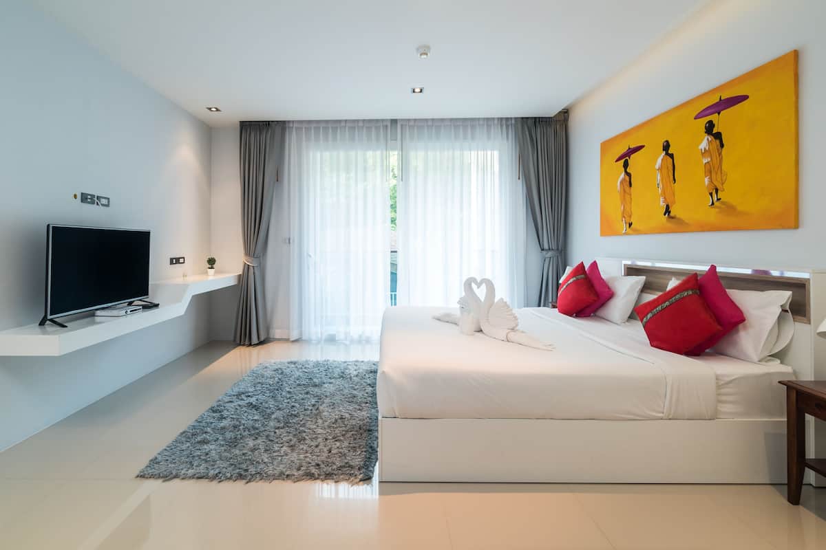 Une chambre d’hôtel moderne dotée d’une télévision à écran plat, d’un grand lit avec des oreillers rouges et blancs, d’un tableau au mur et d’une fenêtre ornée de rideaux légers.