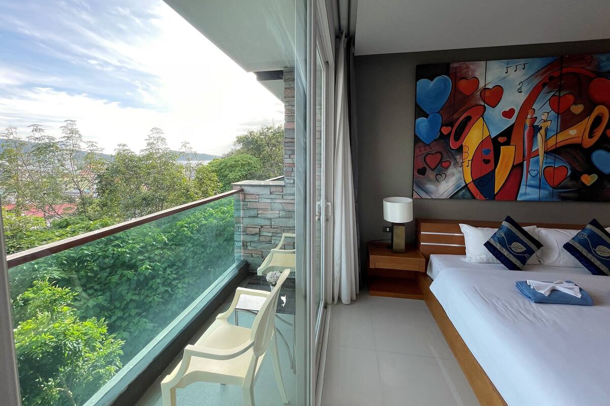 Un balcon avec une balustrade en verre, un lit avec une literie blanche, une table de chevet avec une lampe, et une peinture abstraite aux couleurs vives accrochée au mur.
