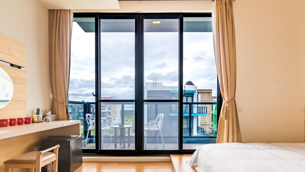 A hotel room with a bed, a desk with a chair, a small refrigerator, and a view of the city through large windows. - Chen Feng Homestay (Magong)
