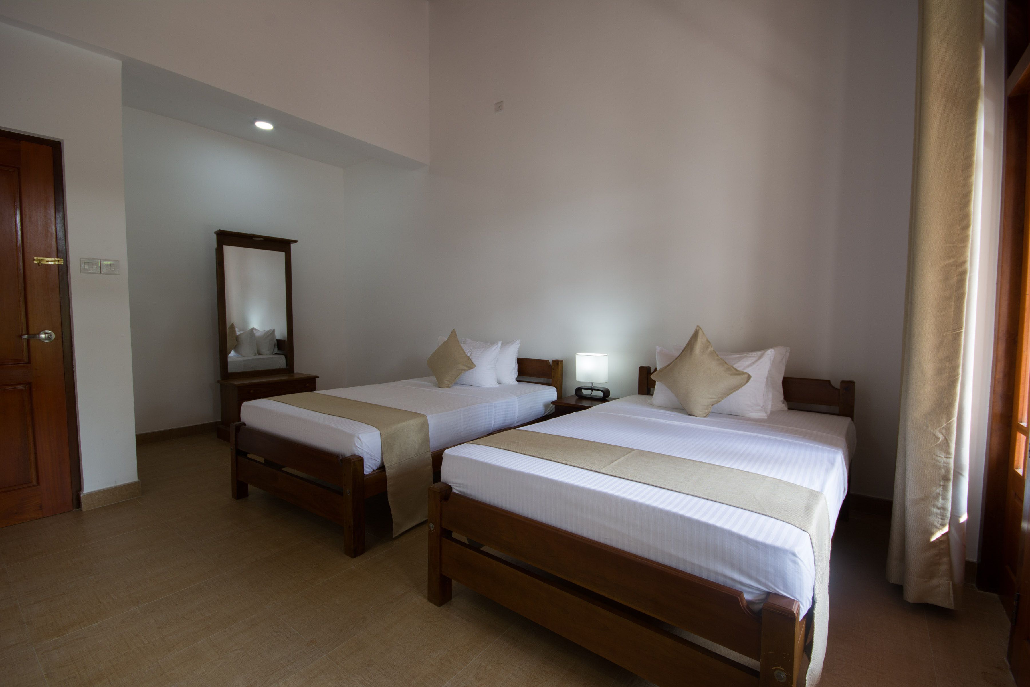 A hotel room with two single beds, a wooden floor, a mirror, a nightstand with a lamp, and a door.