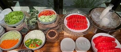Ein Buffet mit verschiedenen Speisen, darunter geschnittenes Gemüse und Obst, sowie einem Schild mit thailändischem Text.