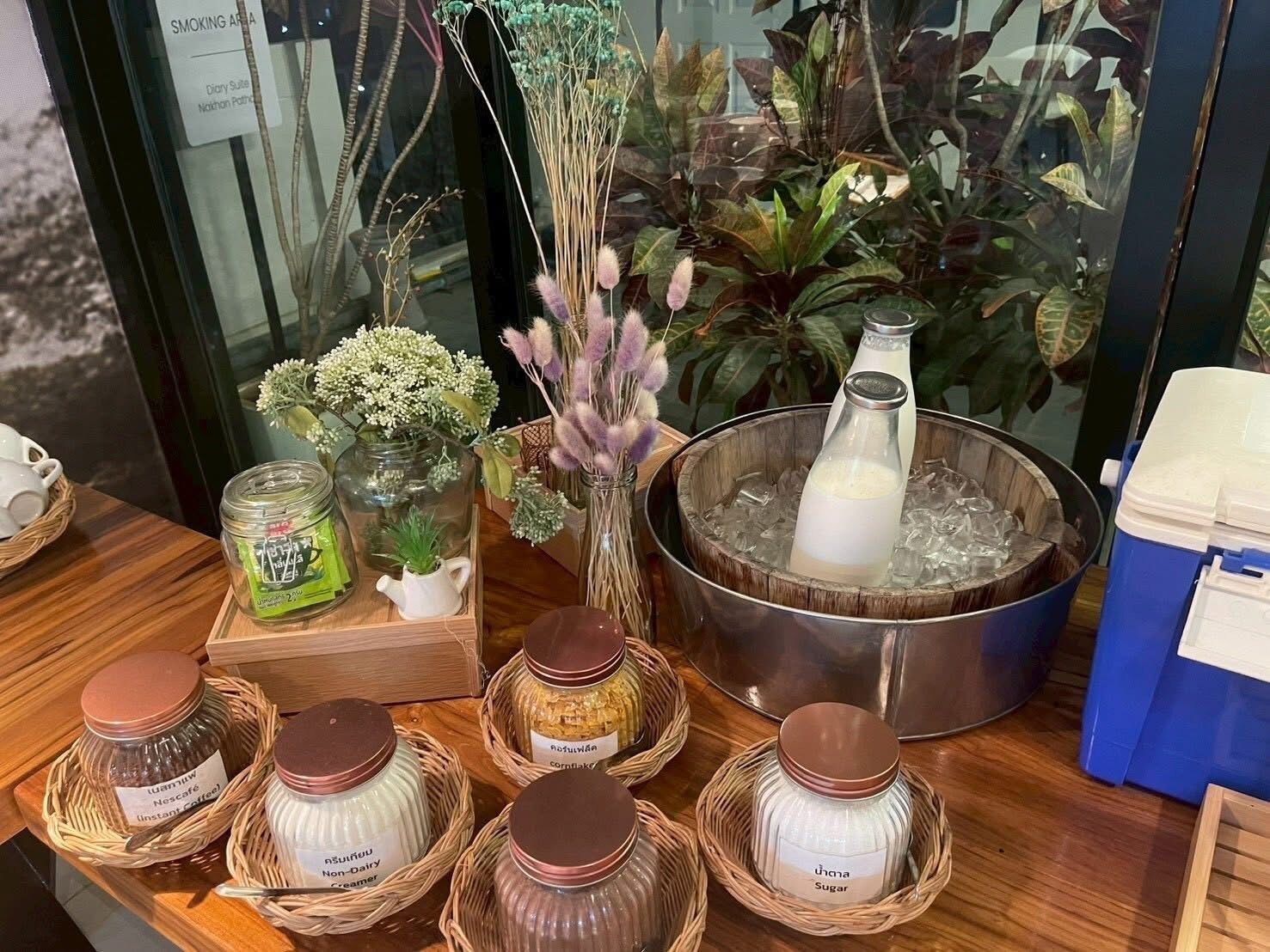 Una exhibición de diversos alimentos y bebidas, incluyendo botes de café instantáneo, crema vegetal, azúcar, así como una botella de leche en un recipiente metálico con hielo.