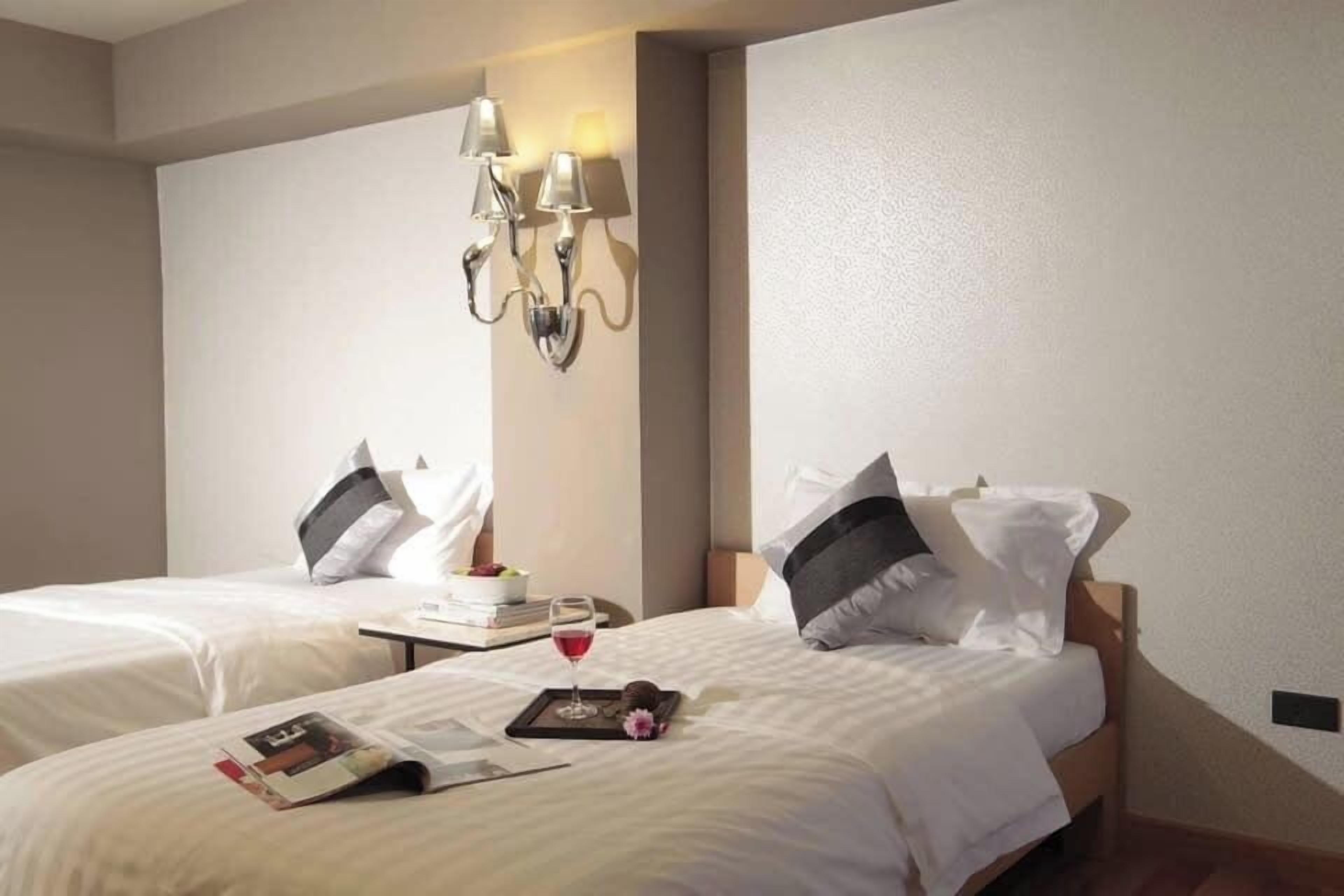 A hotel room with two beds, a wall-mounted lamp, and a tray with a glass of wine and a magazine.