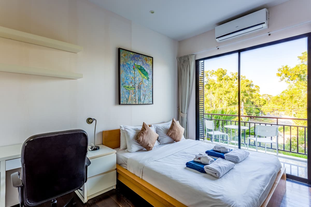 A hotel room with a bed, a desk, a chair, and a large window with a view of greenery.
