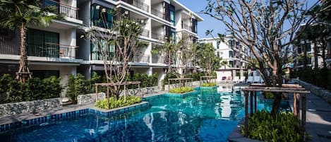 Ett modernt hotell med en swimmingpool omgiven av frodig grönska och en klar, blå himmel.
