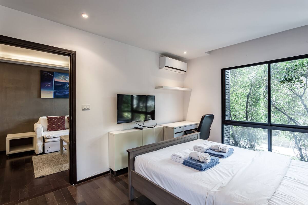 A modern hotel room with a large bed, a desk with a flat-screen TV, a chair, and a window with a view of trees.