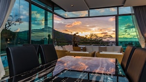 Un comedor moderno con una mesa de vidrio, muebles de mimbre y una vista panorámica del atardecer sobre montañas.
