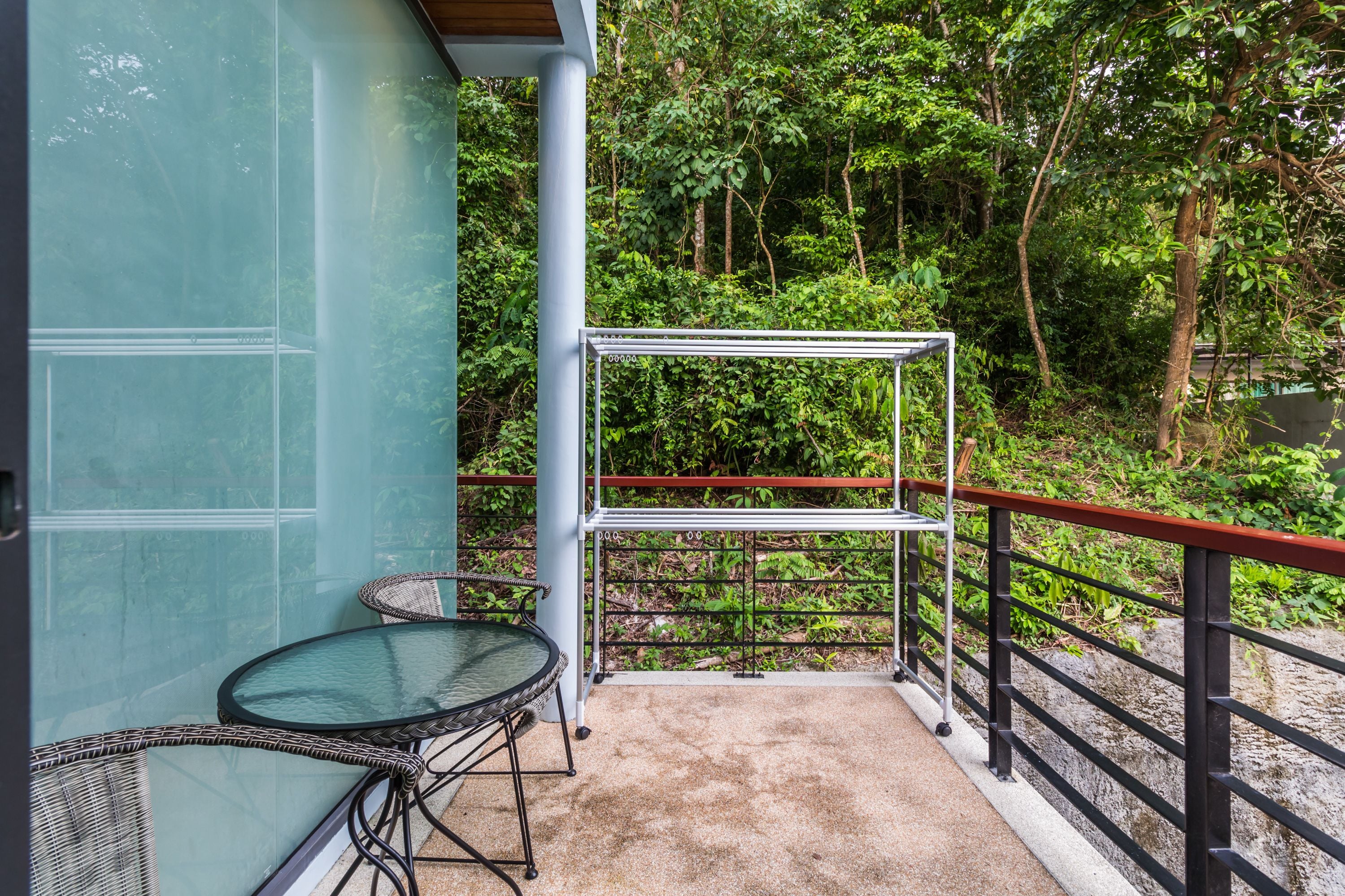 Un balcon agrémenté d’une table en verre et de chaises en osier, entouré de verdure et muni d’une rambarde en métal.