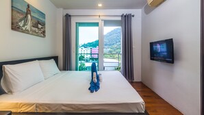 A hotel room with a bed, a painting on the wall, a television, and a view of buildings and hills outside the window. - Heaven Lagoon Apartments by Lofty (Patong)