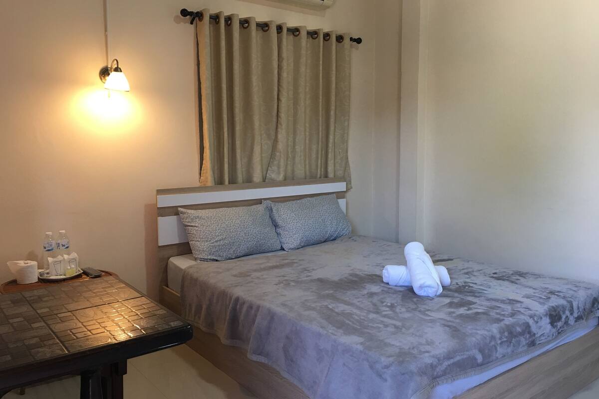 A neatly made bed with a grey blanket and two pillows, a small table with a bottle and glasses, and a wall-mounted air conditioner.