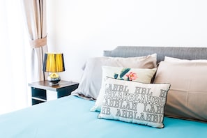 Una cama bien tendida con una colcha azul, una almohada blanca con diseño floral y texto, y una lámpara sobre una mesita de noche.