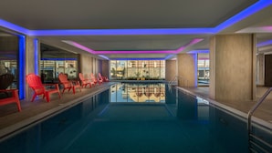 A swimming pool with red chairs and a view of buildings through the windows. - Blue Inn (Abha)