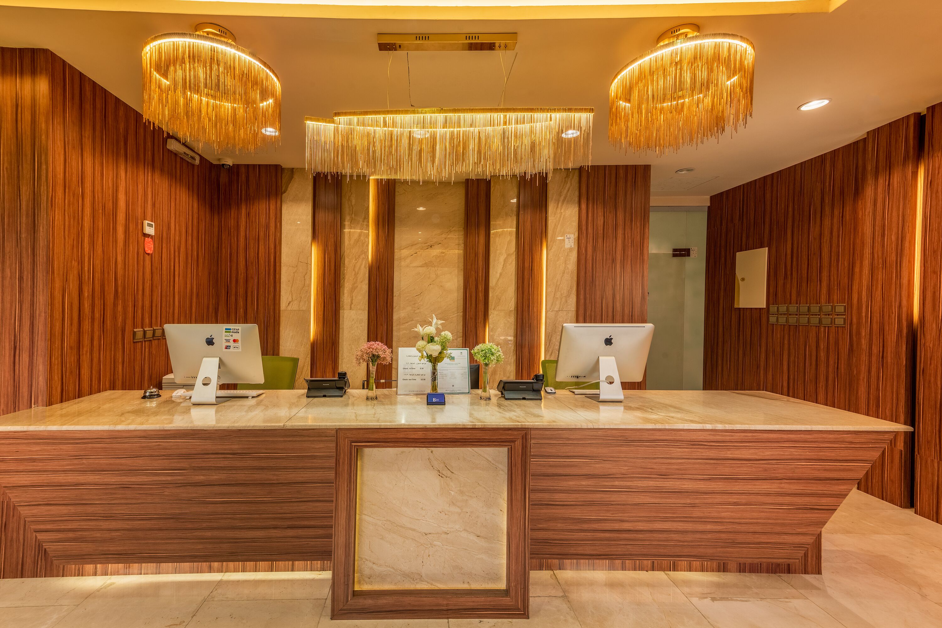 Una recepción con dos computadoras Apple iMac, un panel de pared de madera y una lámpara de araña grande y ornamentada.