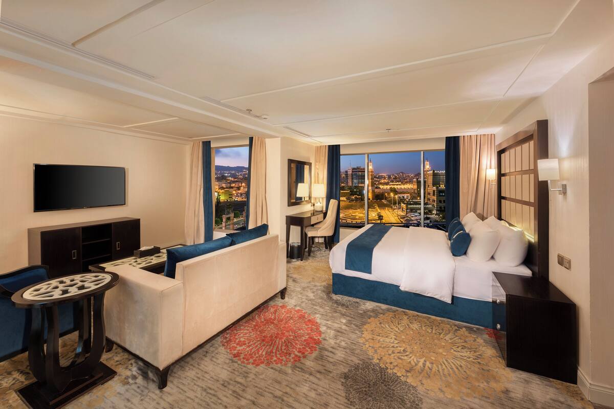 A hotel room with a large bed, a sofa, a desk, and a TV. The room has a city view.