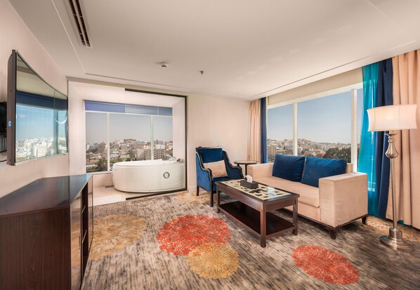 A modern hotel room with a large window offering a city view, a sofa, a chair, a coffee table, and a TV on a wooden cabinet. - Blue Inn (Abha)