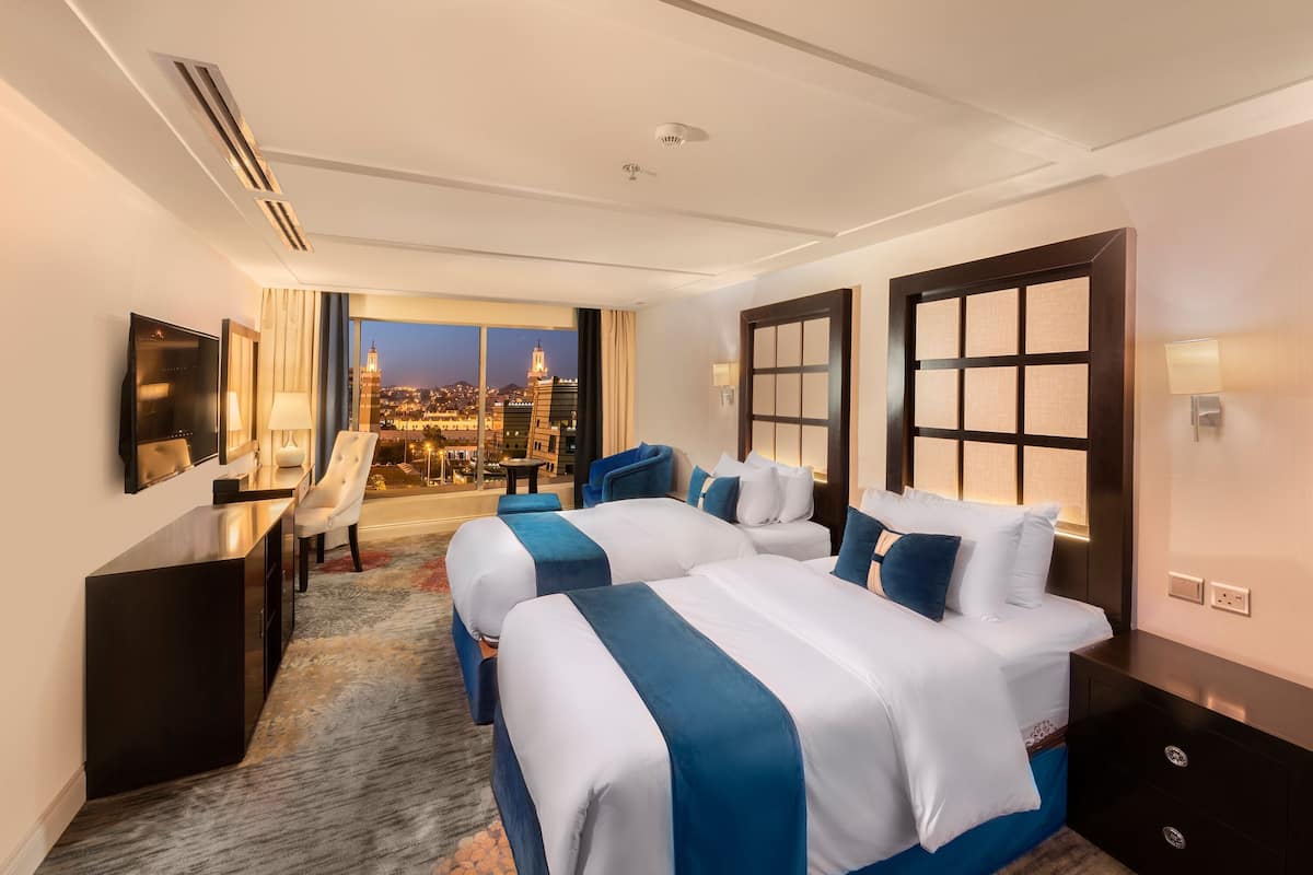 A hotel room with two beds, a desk, a TV, and a view of a cityscape.