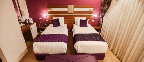 Habitación de hotel con dos camas, cabecero de madera, un reposapiés morado y una ventana con cortinas.