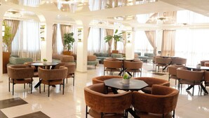 A spacious hotel lobby with multiple seating areas, including round tables and sofas, and large windows with curtains.
