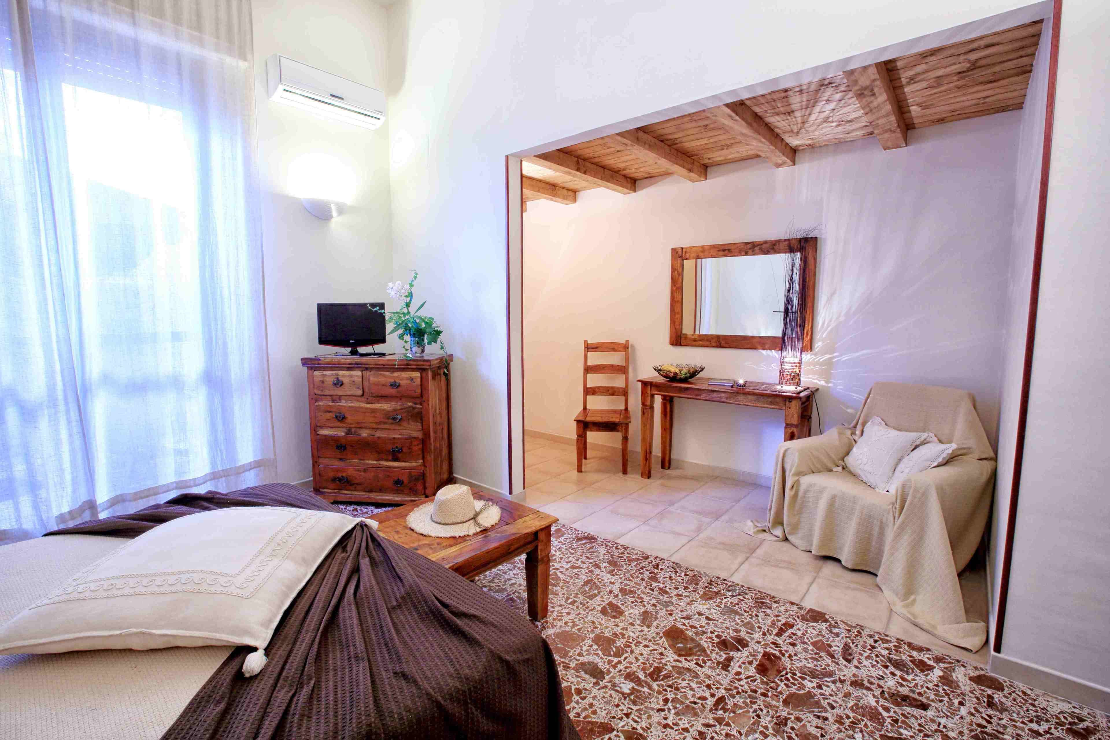 A bedroom with a bed, a wooden dresser, a desk with a chair, and a bathroom with a chair and a mirror.