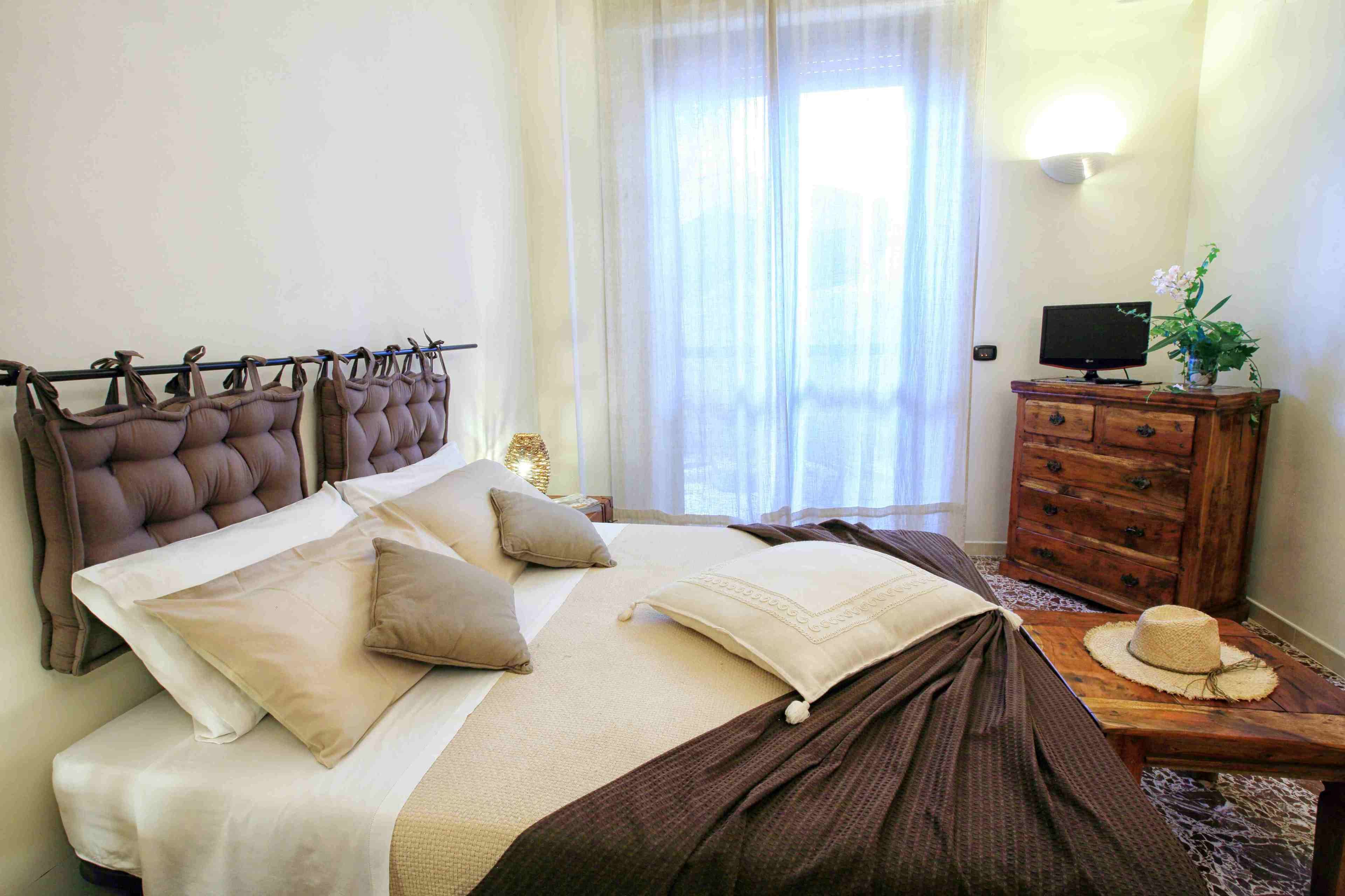 A neatly made bed with a brown headboard, a wooden dresser with a television, and a window with sheer curtains.