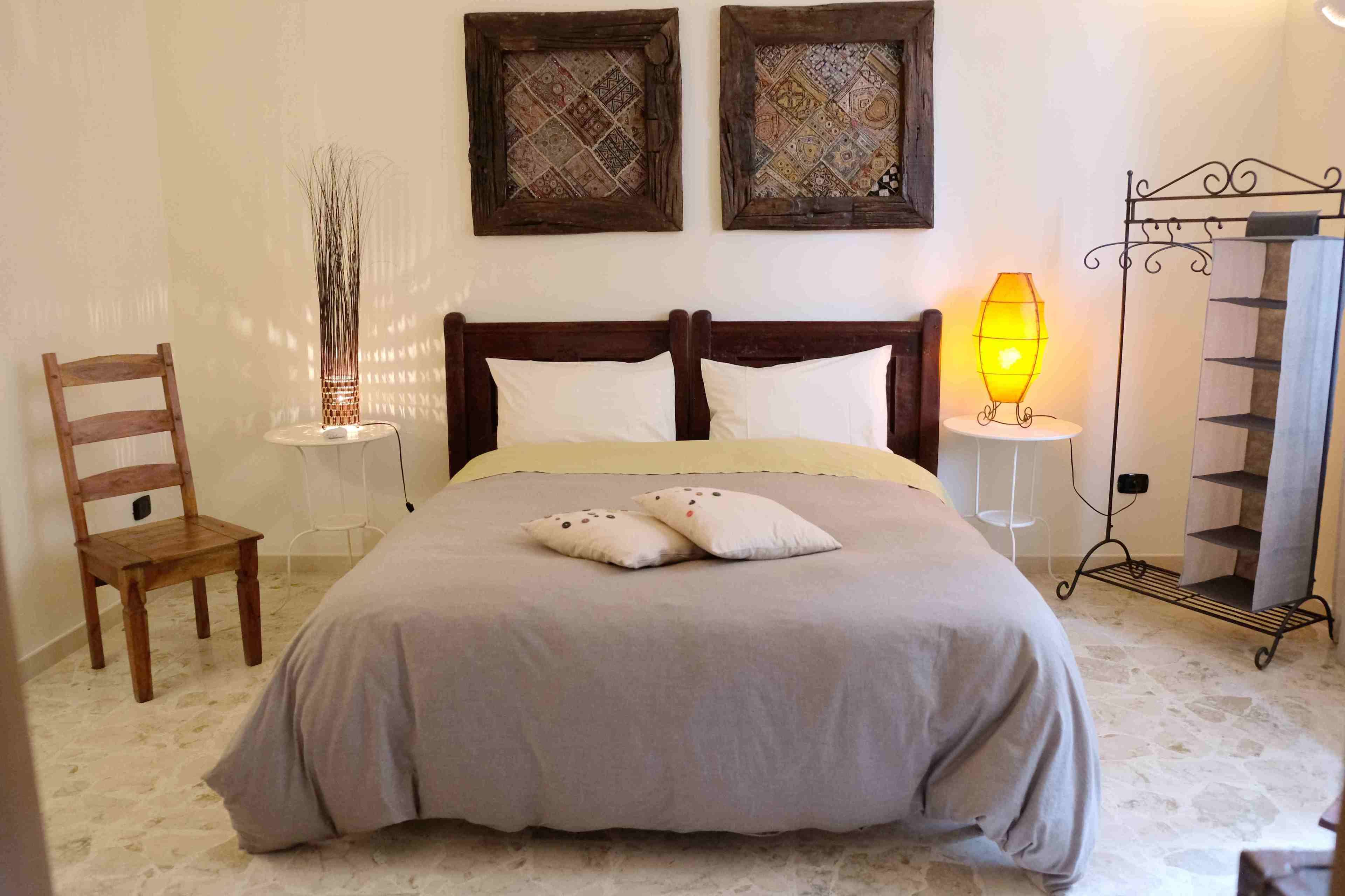 Quarto com cama, duas molduras na parede, abajur, cadeira de madeira e estante de metal.