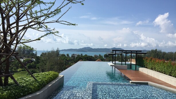 A rectangular infinity pool with clear blue water, surrounded by a wooden deck and a low stone wall. In the background, there is a view of a coastal landscape with mountains and a clear sky. - 6 Avenue Surin by Lofty (Choeng Thale)