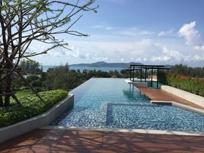 A rectangular infinity pool with clear blue water, surrounded by a wooden deck and a low stone wall. In the background, there is a view of a coastal landscape with mountains and a clear sky.