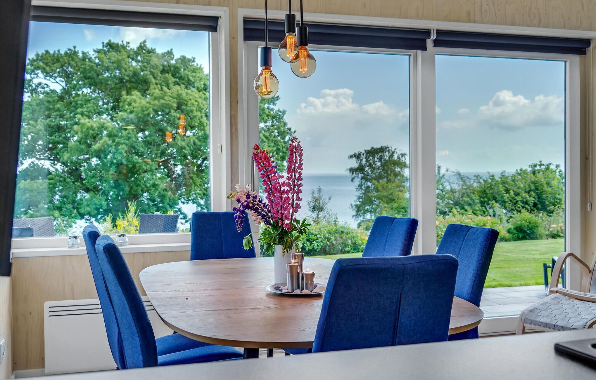Comedor con mesa redonda de madera, sillas tapizadas en azul y vistas a zonas verdes y agua a través de grandes ventanales.