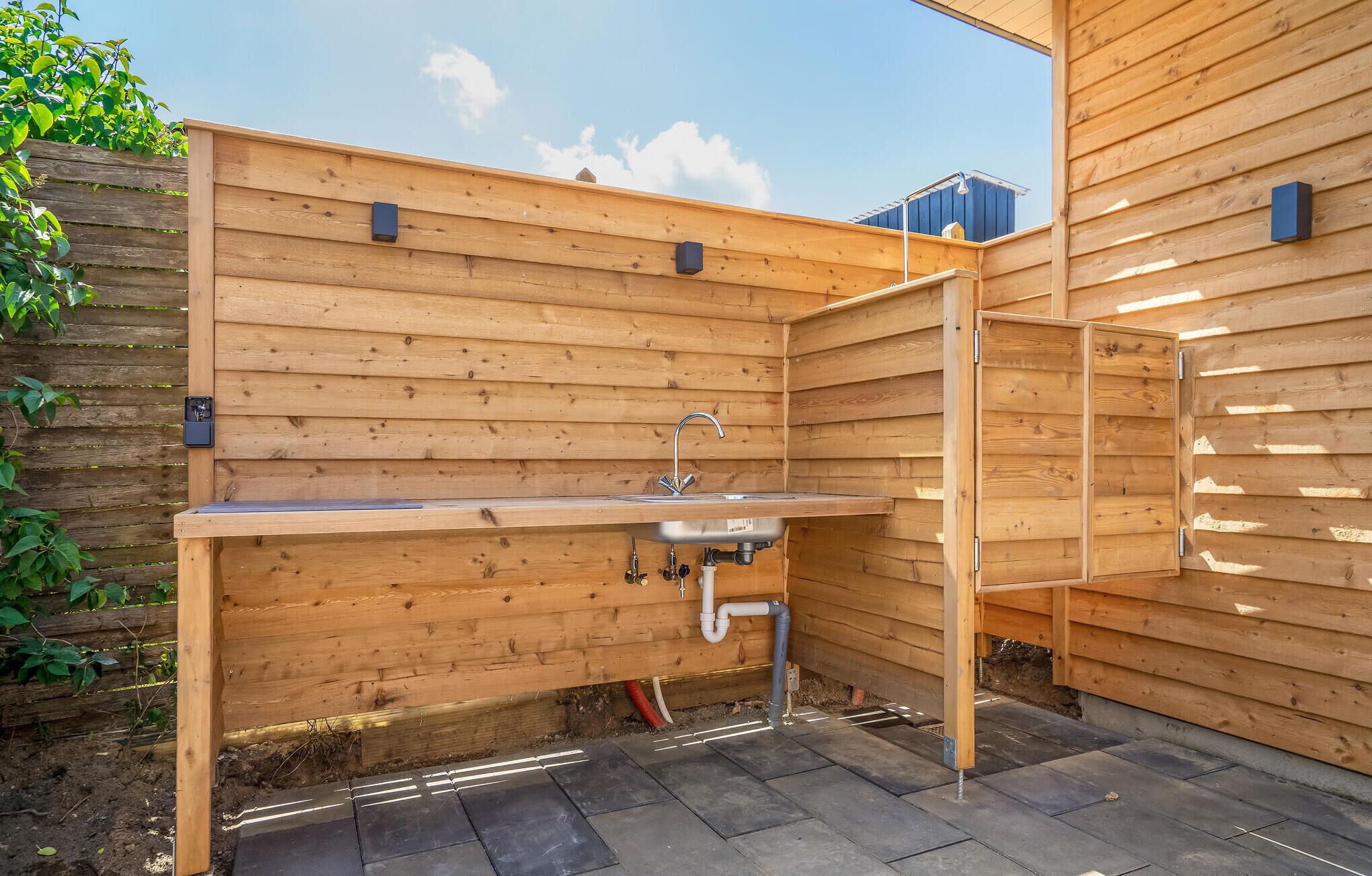 A wooden outdoor kitchen with a sink and built-in cabinets, surrounded by a wooden fence and a paved patio.