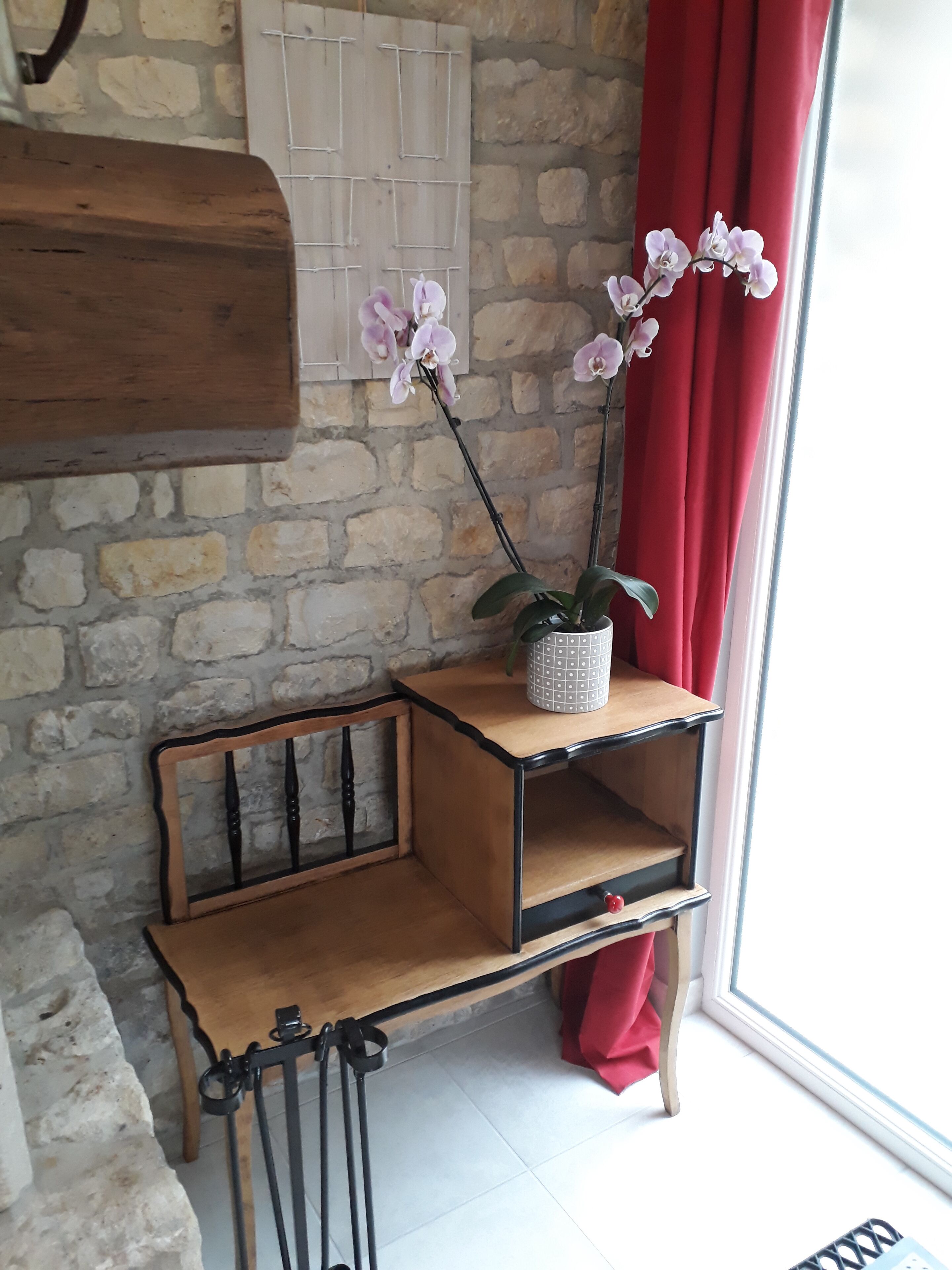 Een kamer met een stenen muur, een houten bankje, een kleine tafel met een potorkidee en een raam met rode gordijnen.