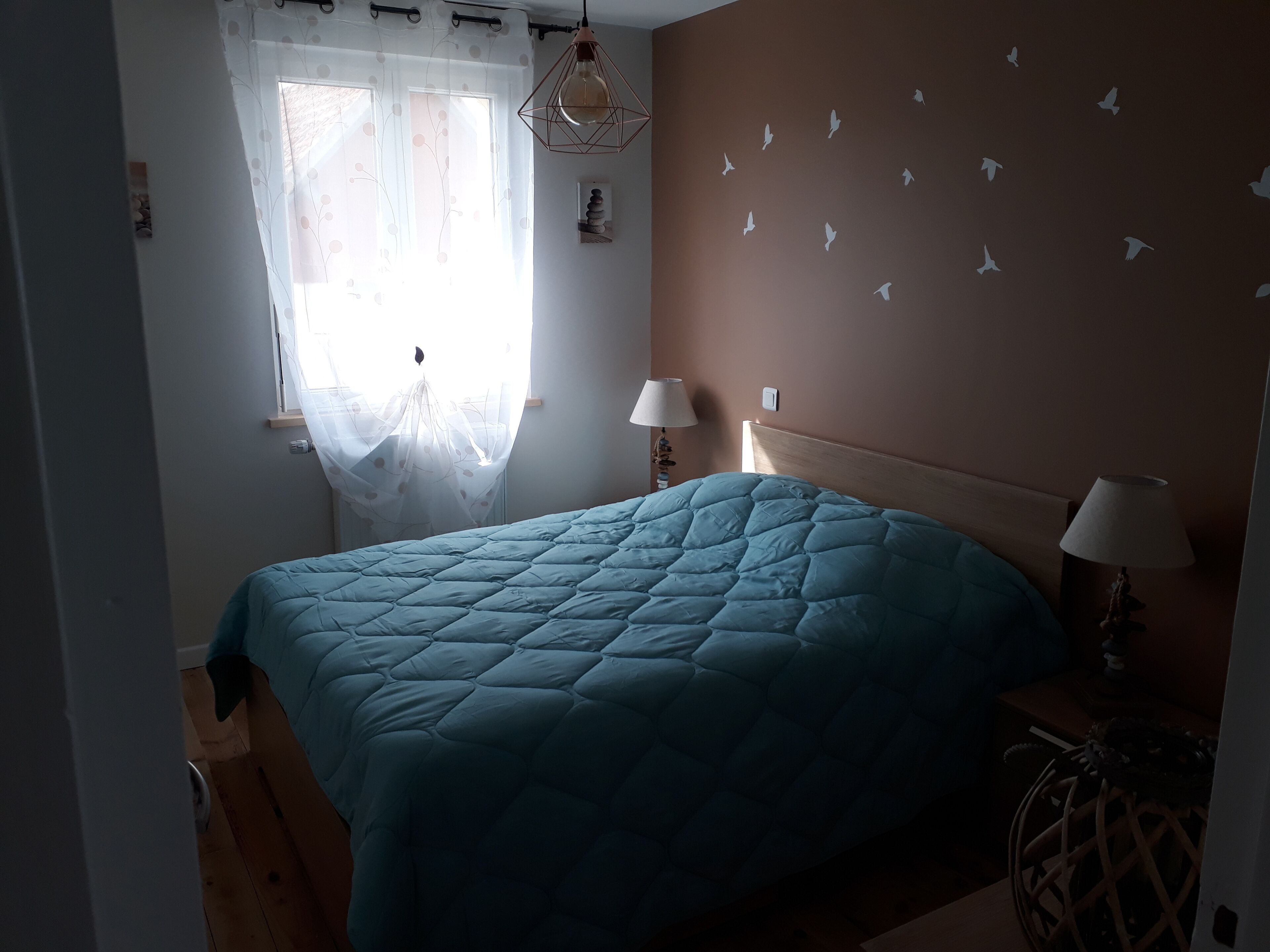 Une chambre à coucher avec un grand lit, deux lampes de chevet, une fenêtre avec des rideaux légers et un mur décoré d’autocollants représentant des oiseaux.