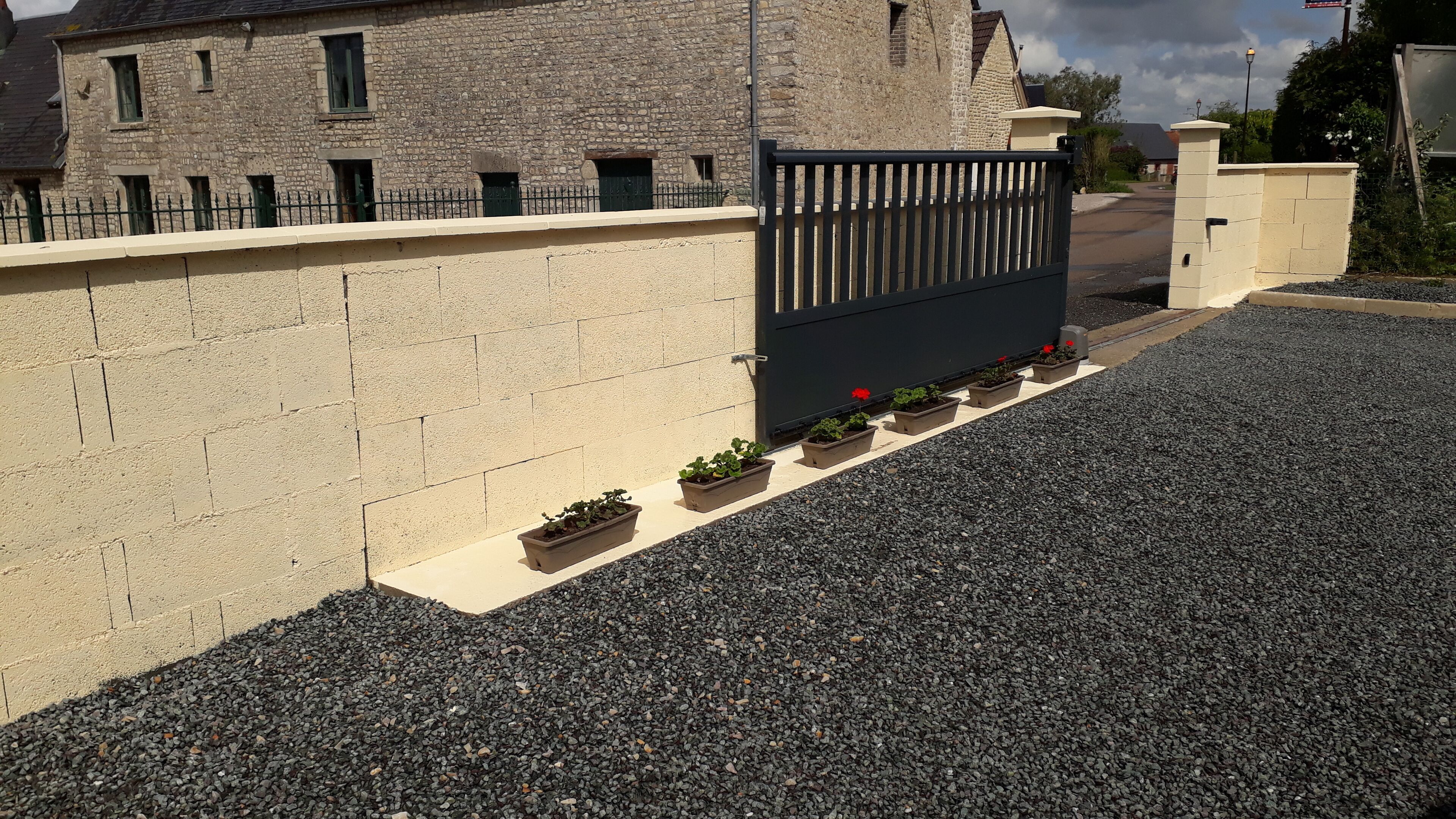 A stone building with a gated entrance, a gravel driveway, and potted plants along the wall.