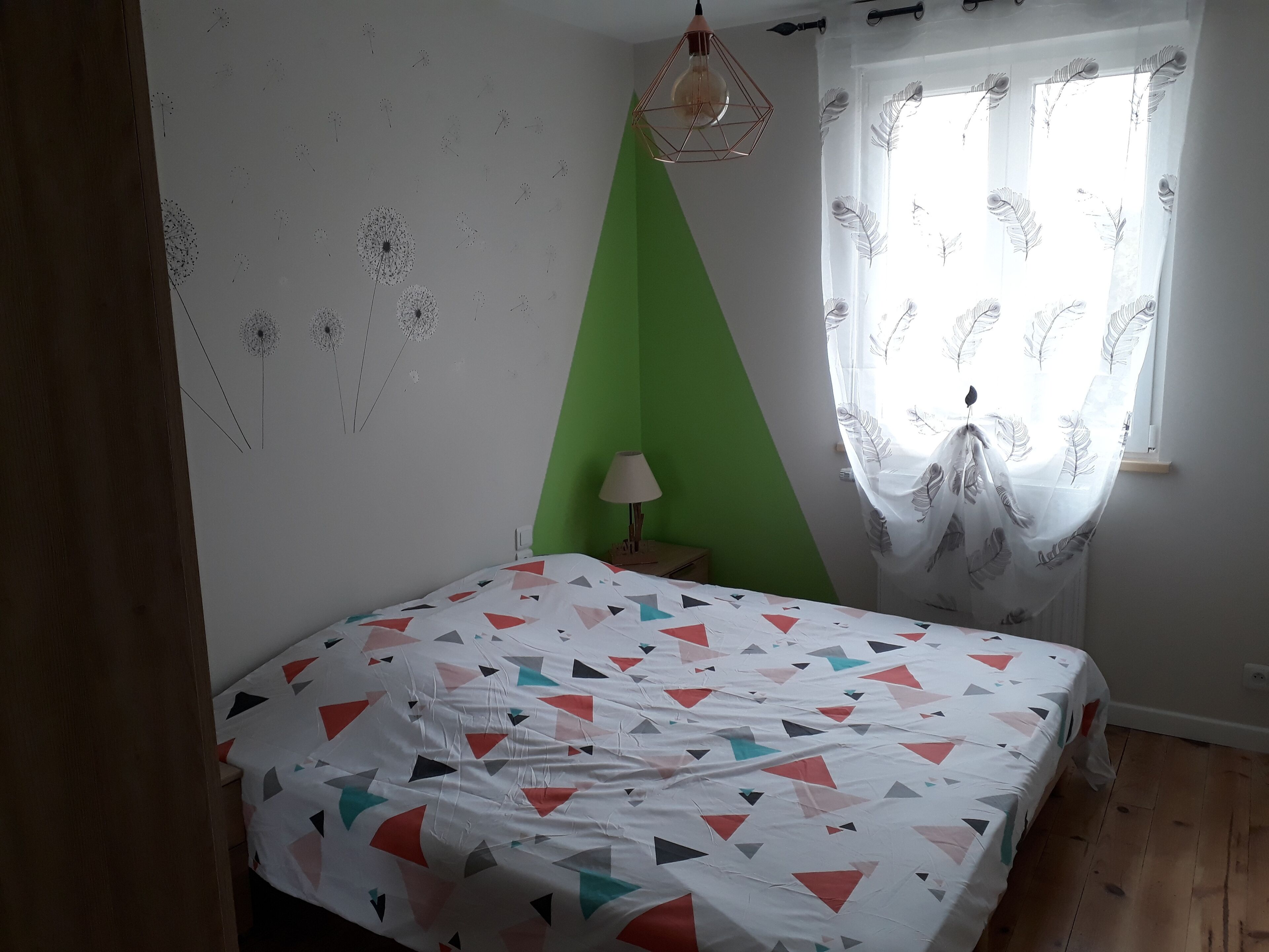 Un dormitorio con cama, una pared de acento verde, una ventana con cortinas translúcidas y una lámpara colgante.
