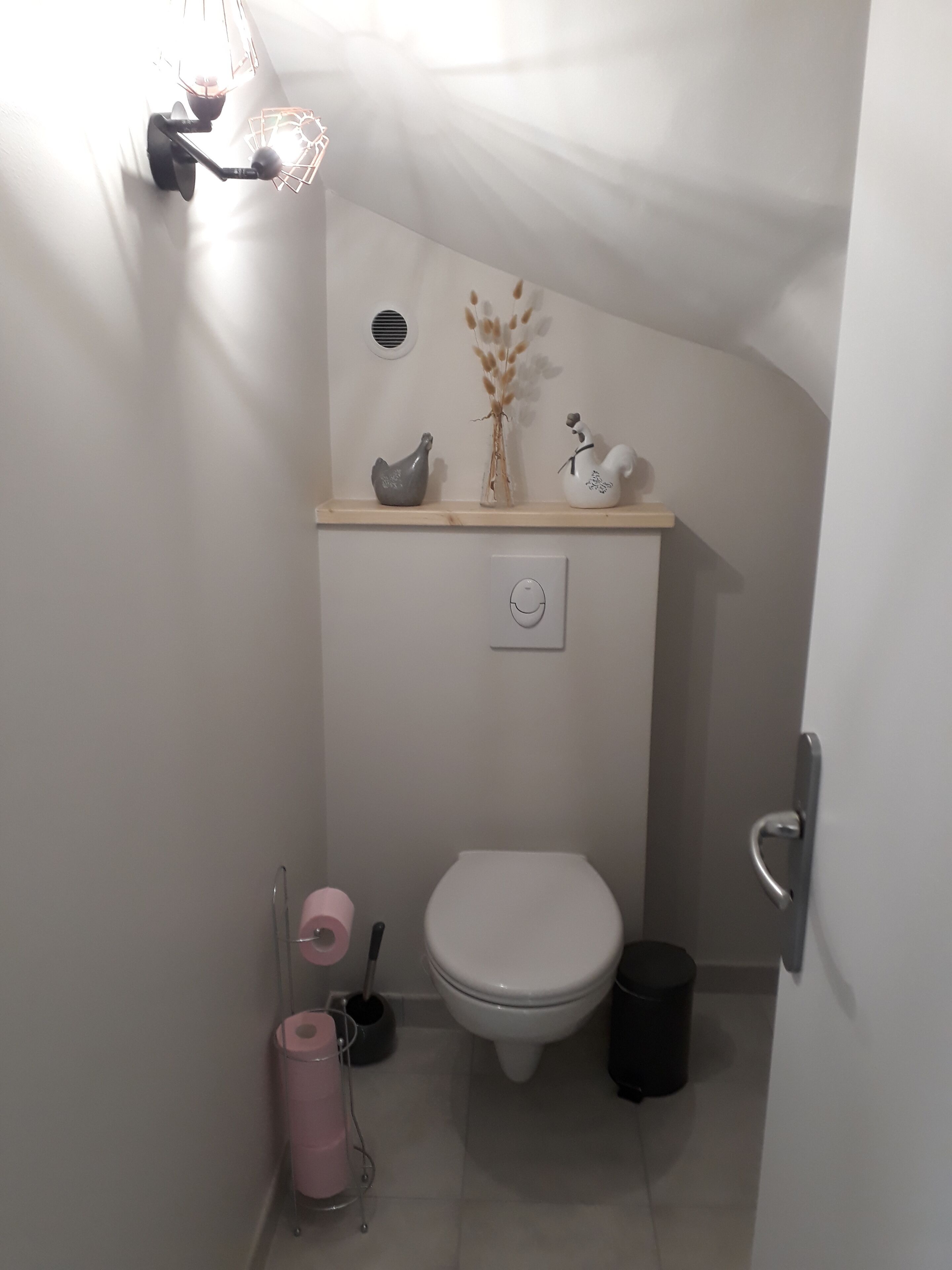 A small, compact bathroom with a toilet, a sink, and a shelf above the toilet holding decorative items.