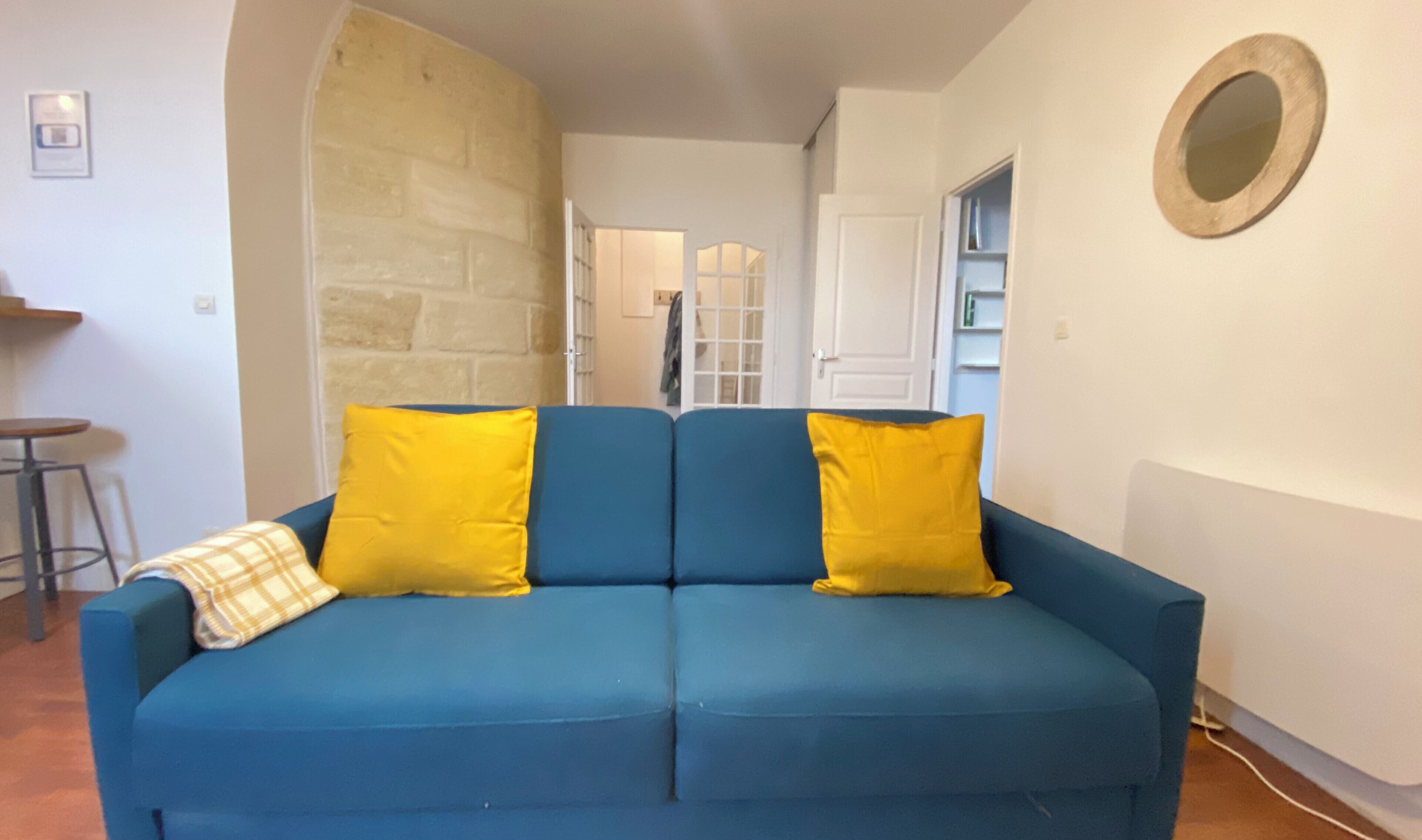 Un sofá azul con cojines amarillos en una habitación con pared de piedra, un espejo redondo en la pared, y una puerta que da a otra habitación.