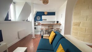 Un salon moderne avec un canapé bleu, des coussins jaunes, une table basse blanche et un coin cuisine doté d’un mur d’accent bleu, d’une horloge et de tabourets de bar.