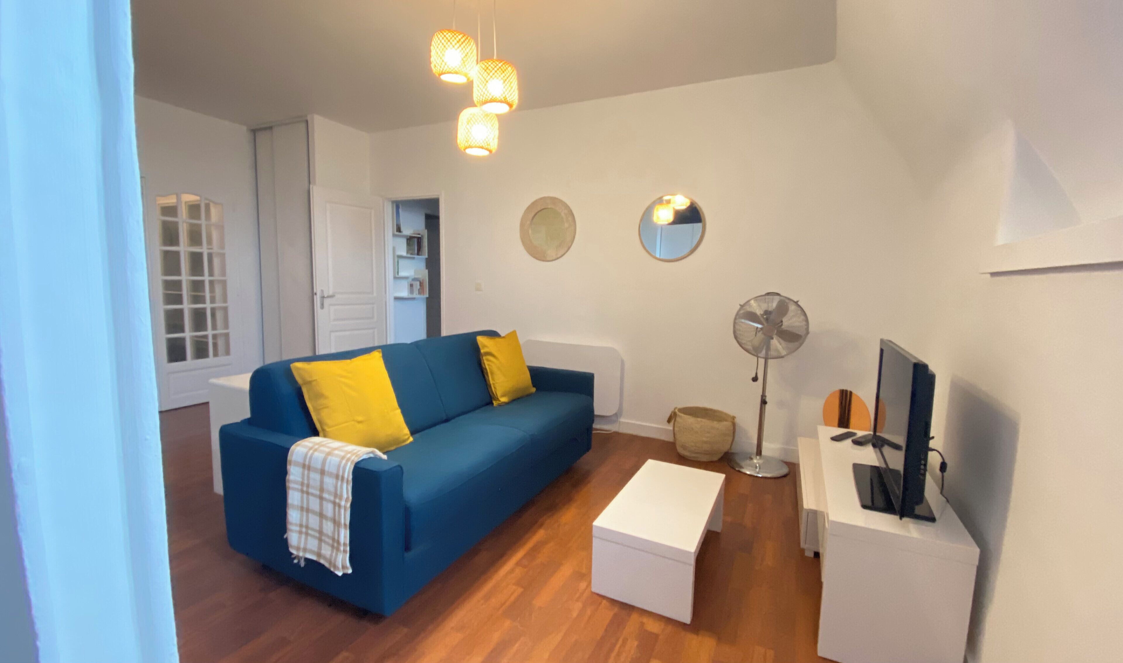 A living room with a blue sofa, yellow pillows, a white side table, a standing fan, and a television.