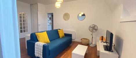 A living room with a blue sofa, yellow pillows, a white side table, a standing fan, and a television.