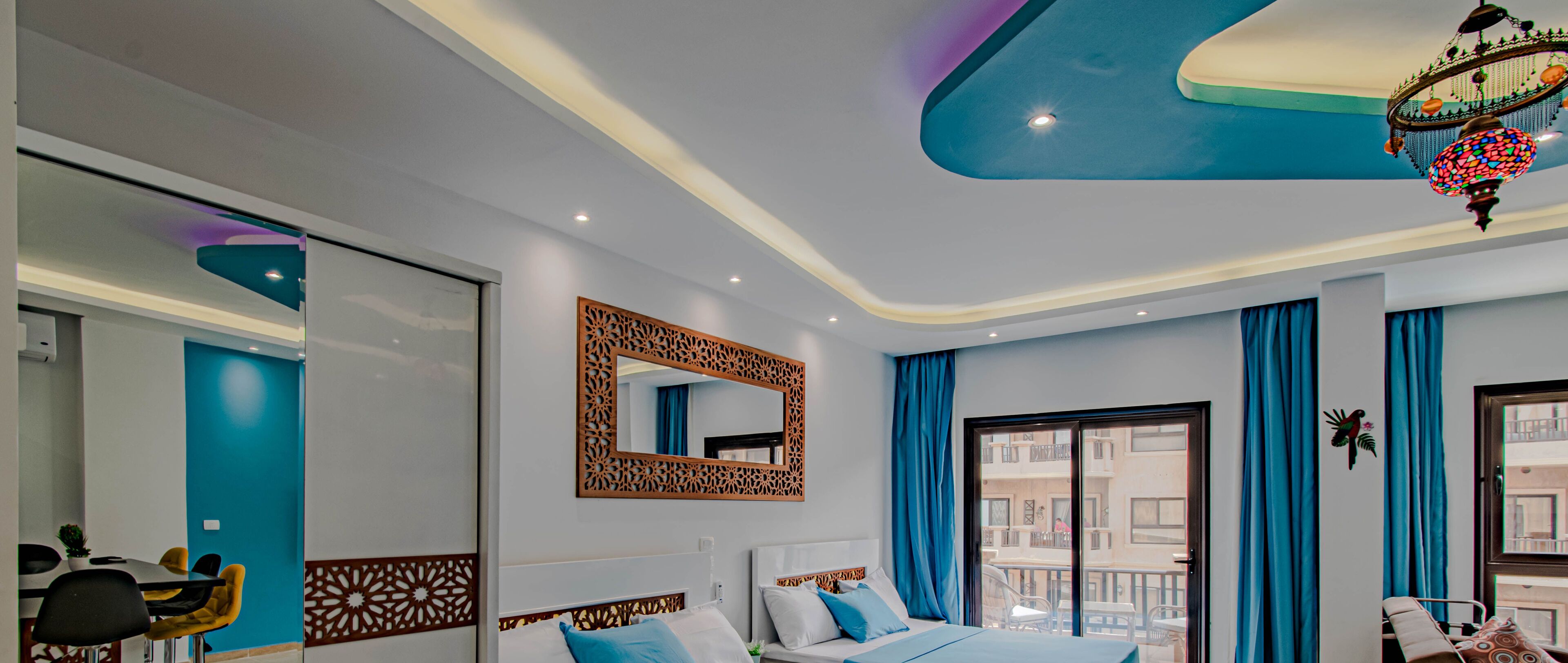 A modern hotel room with a large bed, a balcony with blue curtains, and a decorative chandelier.