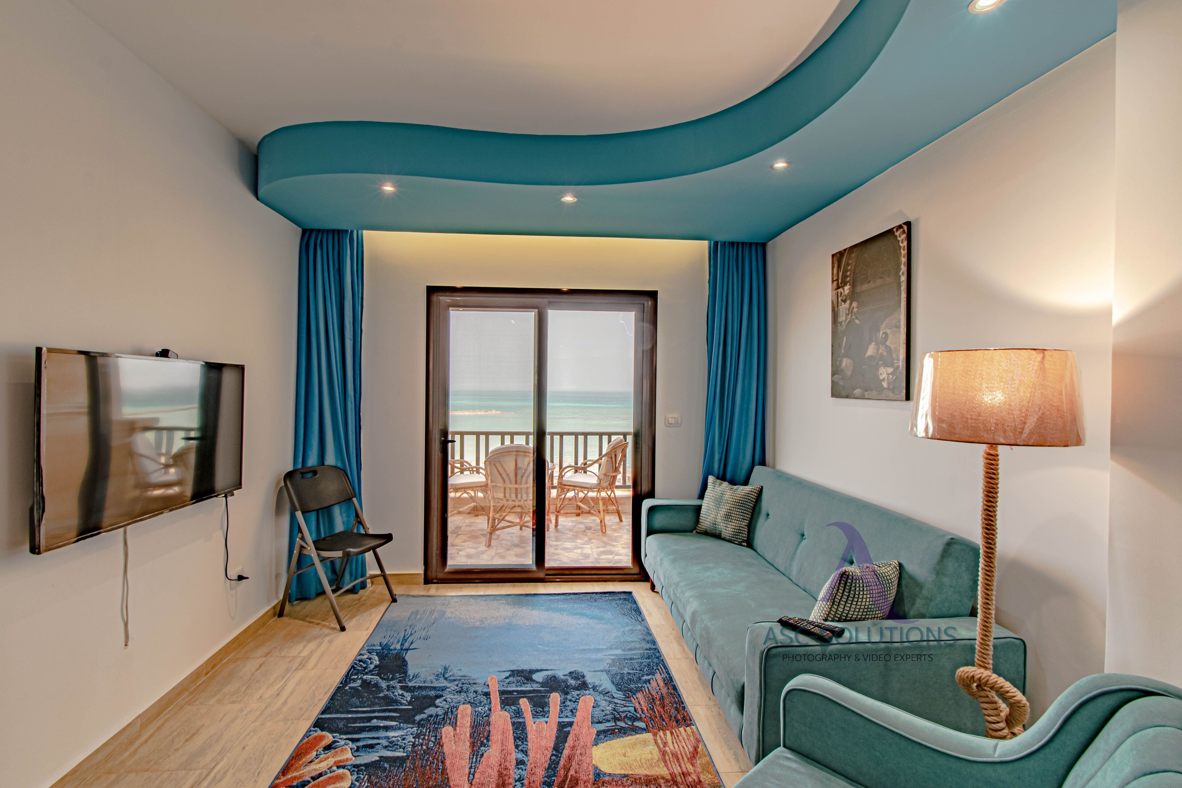 Una sala de estar con sofá, televisión, sillón, lámpara y vista a la playa a través de un ventanal.